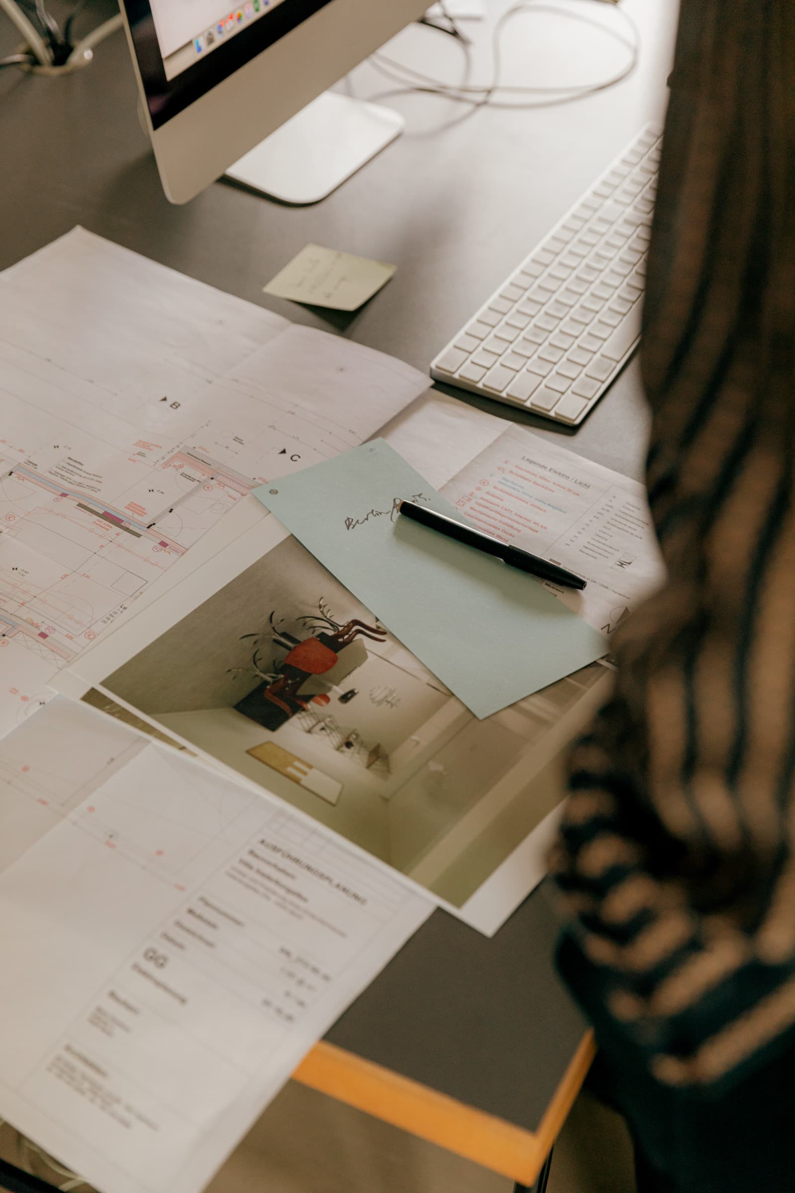 Planos de planta, foto impresa de un interior y bolígrafo extendidos en una mesa junto al teclado, vistos por encima del hombro de alguien