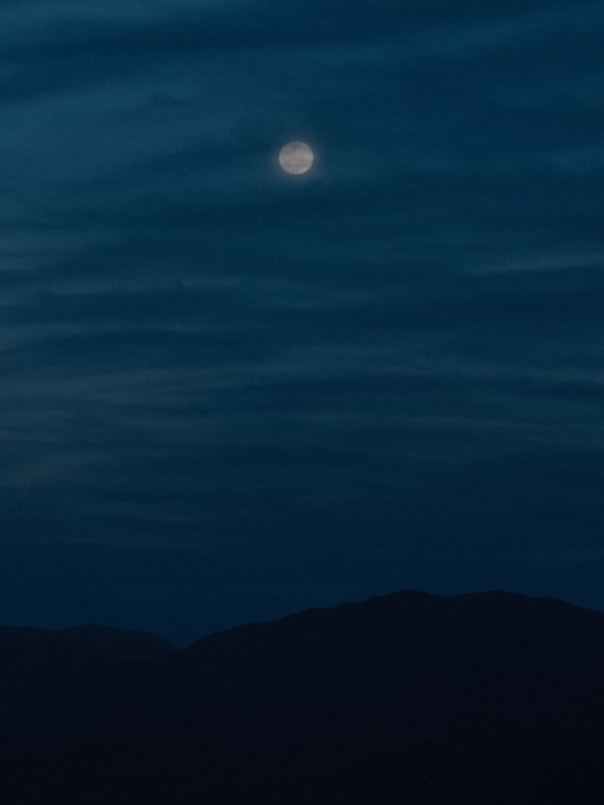Luna llena brillando a través de finas franjas de nubes, muy por encima de la silueta de una cresta montañosa